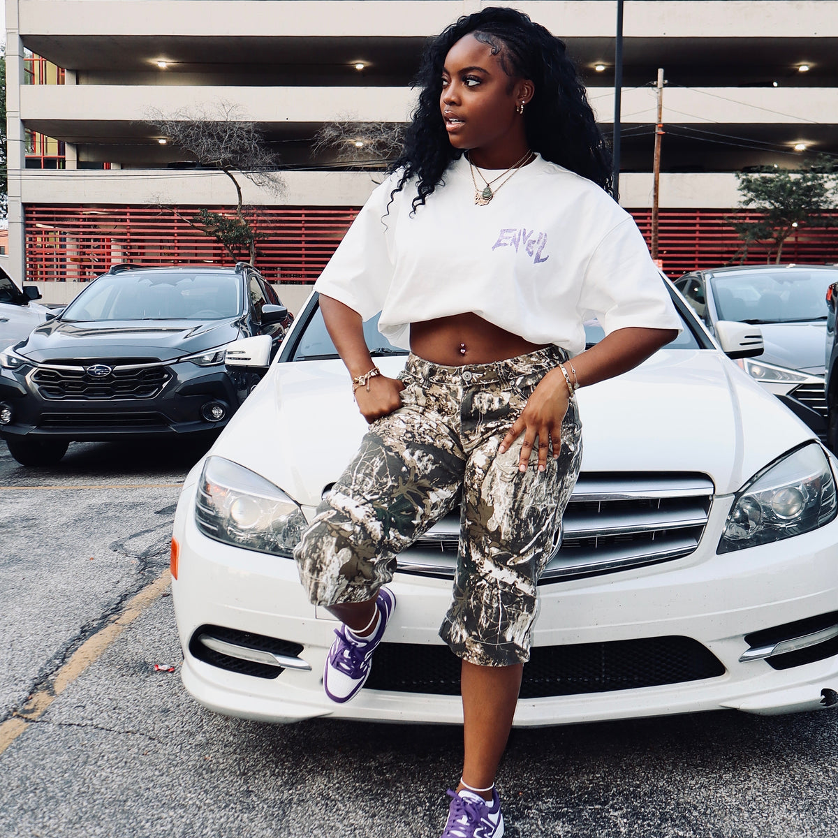 A woman posing in front of a white car, wearing a white cropped t-shirt with purple text, camouflage-patterned cropped pants, and purple sneakers.