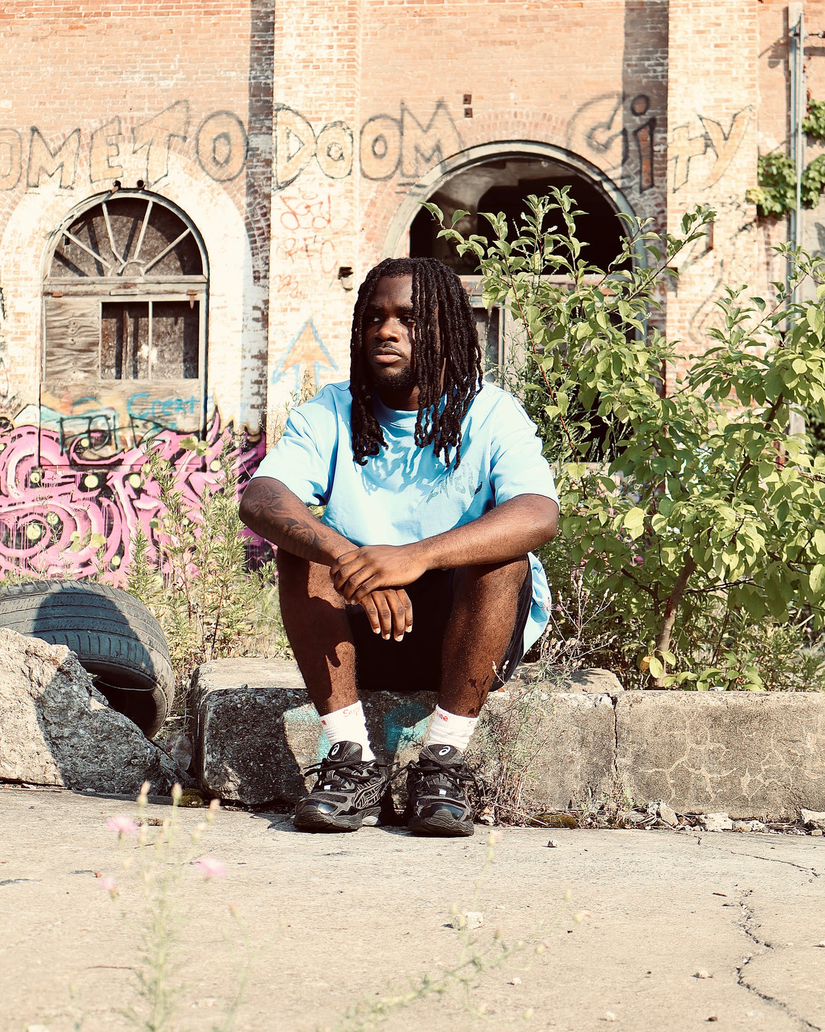 A man with dreadlocks sits on a concrete ledge in front of a brick building with graffiti, wearing a light blue t-shirt, dark shorts, white socks, and black sneakers.